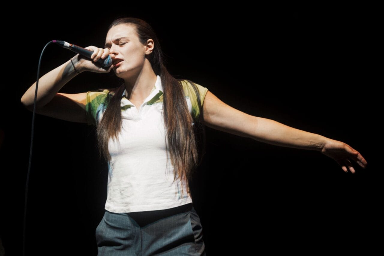 A person with long brown hair sings into a microphone on stage, eyes closed, one arm extended outward as they perform with Common Tongue against a black background.