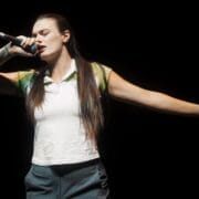A person with long brown hair sings into a microphone on stage, eyes closed, one arm extended outward as they perform with Common Tongue against a black background.