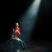 A person sits alone on a crate under a spotlight on a dimly lit stage, ready to share their story in the common tongue, with microphones positioned in front of them.
