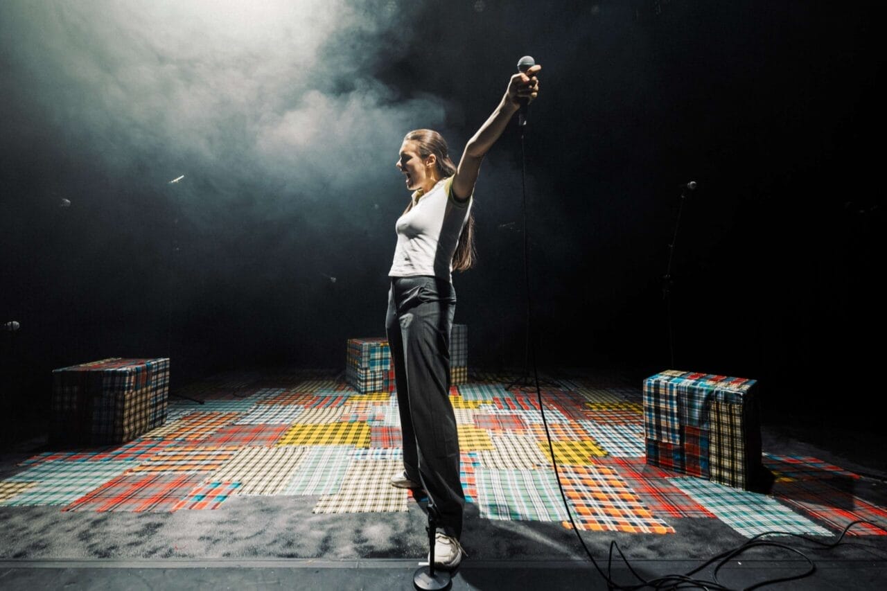 A person stands on a colorful checkered stage, holding a microphone with arms outstretched, ready to speak in the Common Tongue, surrounded by three plaid-patterned block seats.