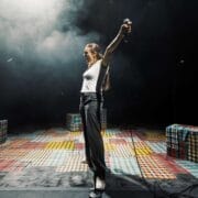 A person stands on a colorful checkered stage, holding a microphone with arms outstretched, ready to speak in the Common Tongue, surrounded by three plaid-patterned block seats.