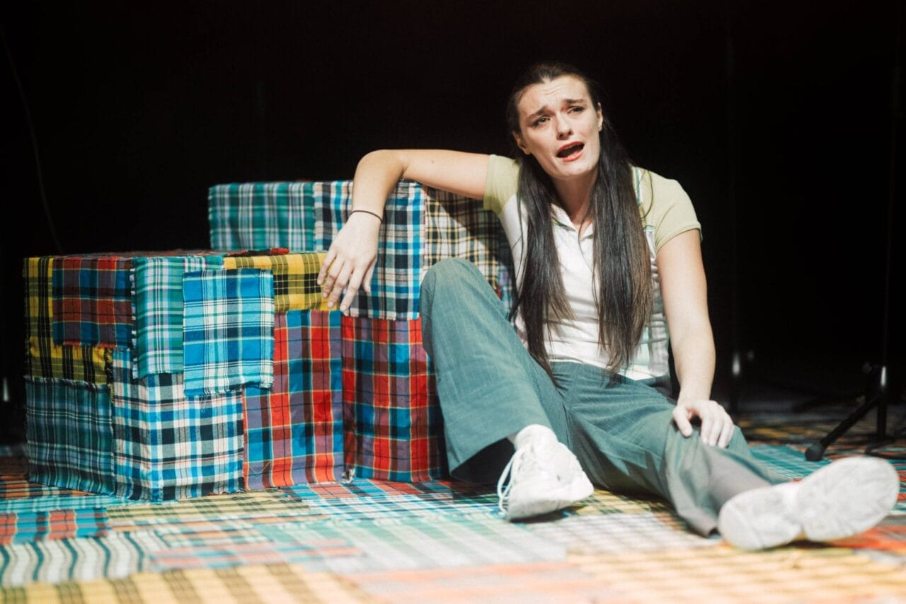 A woman sits on the floor next to colorful plaid-covered boxes, appearing distressed, her mouth open as if searching for words in the Common Tongue, with her arm resting on a box.