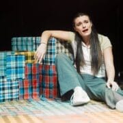 A woman sits on the floor next to colorful plaid-covered boxes, appearing distressed, her mouth open as if searching for words in the Common Tongue, with her arm resting on a box.