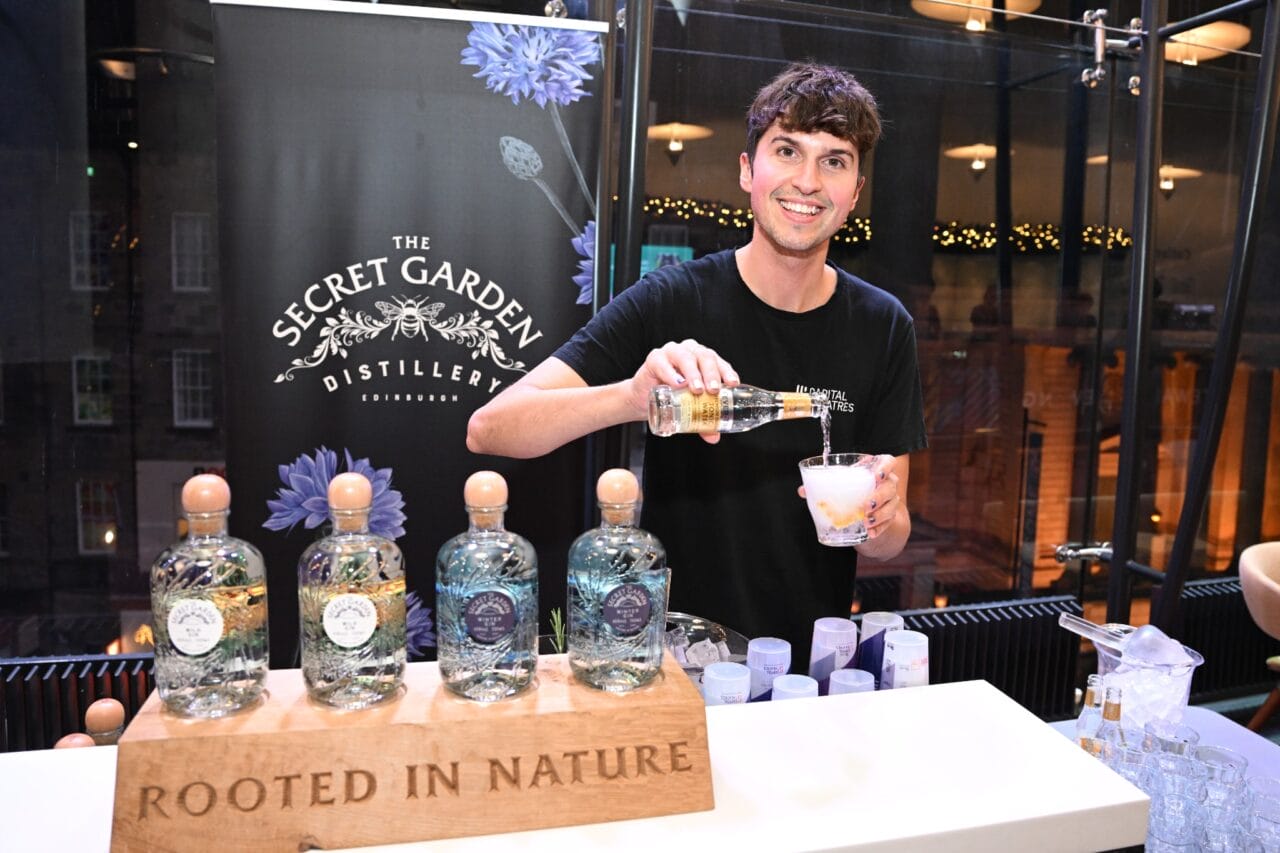 A member of the bar staff pours a drink at a booth featuring bottles labeled “The Secret Garden Distillery” with a sign that reads “Rooted in Nature.”.