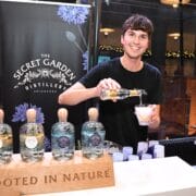 A member of the bar staff pours a drink at a booth featuring bottles labeled “The Secret Garden Distillery” with a sign that reads “Rooted in Nature.”.