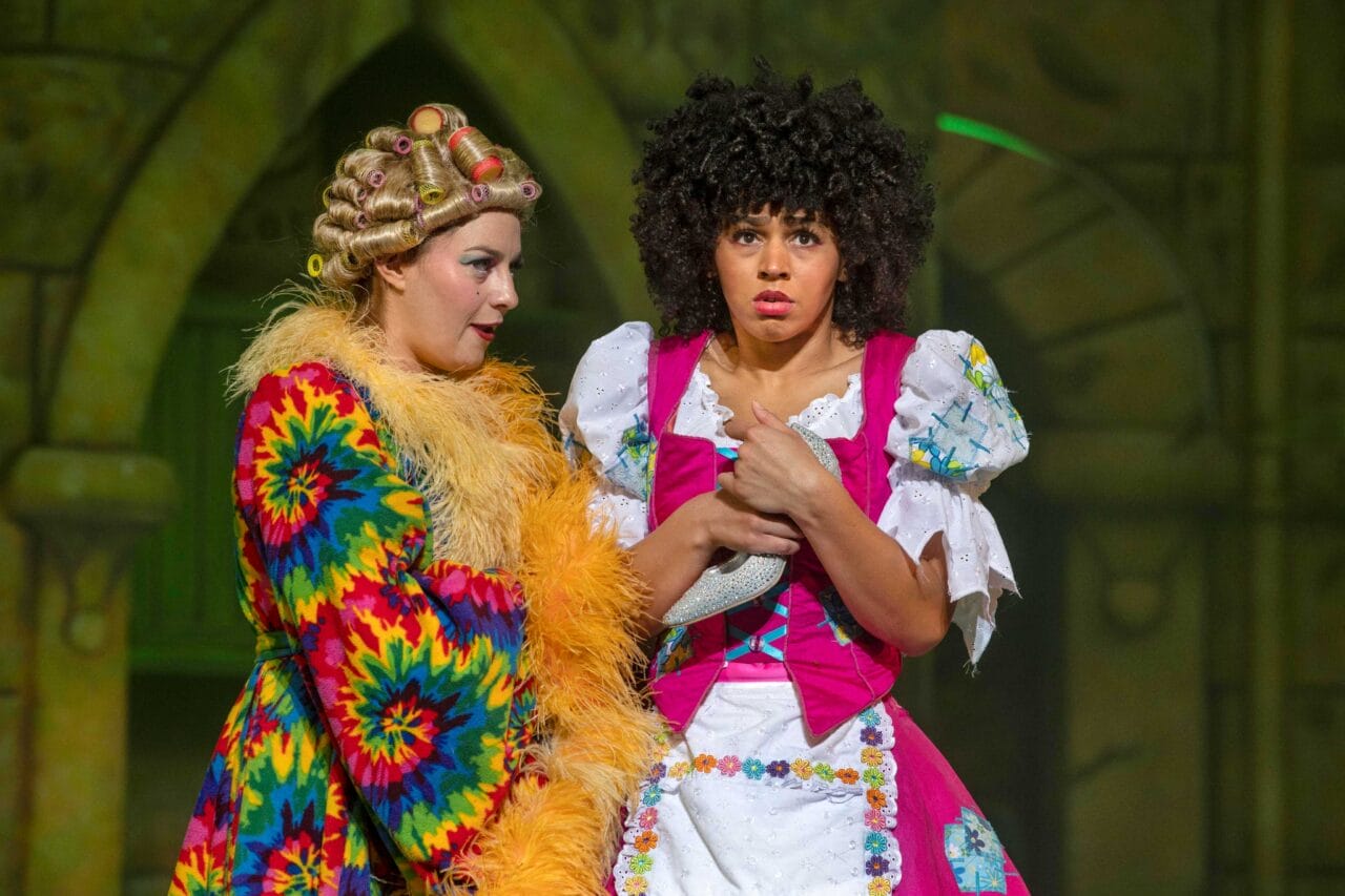 Two actors on stage in colourful costumes; one in a multicoloured robe and curlers, the other in a pink dress looking worried, holding a white cloth.