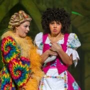 Two actors on stage in colourful costumes; one in a multicoloured robe and curlers, the other in a pink dress looking worried, holding a white cloth.