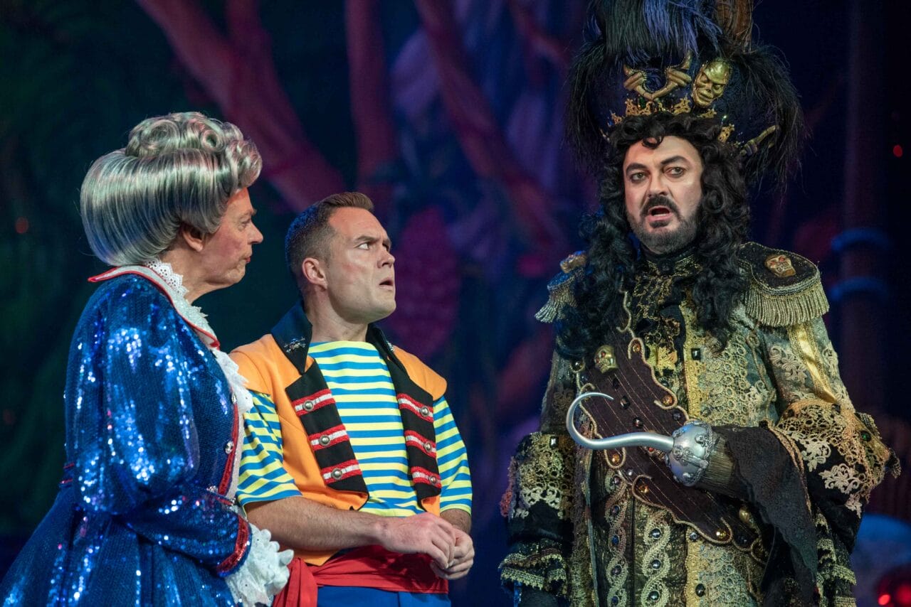 Three actors in colourful, elaborate costumes perform on stage; one is dressed as a pirate with a hook hand, another in a blue dress and wig, and the third in a striped shirt and vest.