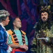 Three actors in colourful, elaborate costumes perform on stage; one is dressed as a pirate with a hook hand, another in a blue dress and wig, and the third in a striped shirt and vest.