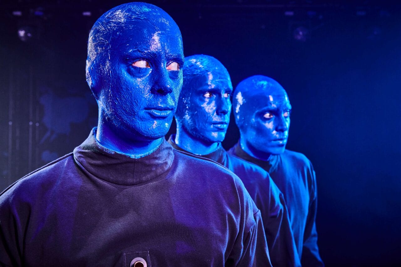 Three people with blue-painted faces and bald heads wearing black outfits stand in a row against a dark background.