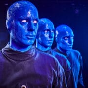 Three people with blue-painted faces and bald heads wearing black outfits stand in a row against a dark background.