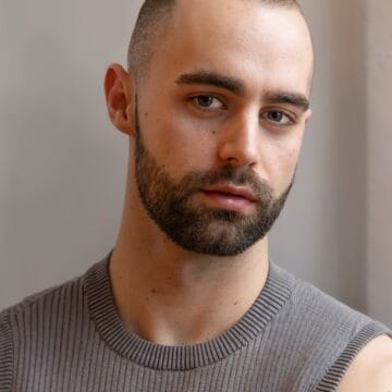 A man with a shaved head and beard, wearing a sleeveless gray ribbed shirt, looks at the camera with a neutral expression against a plain background.