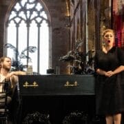 A woman sings beside a coffin and a portrait of an older man in a church, evoking the emotional depth of The Bear; a man sits nearby, watching her intently.
