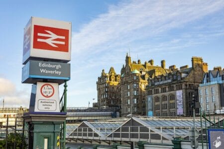 Edinburgh Waverley train station sign in front of historic buildings, with clear blue sky and the City Art Centre visible in the background.