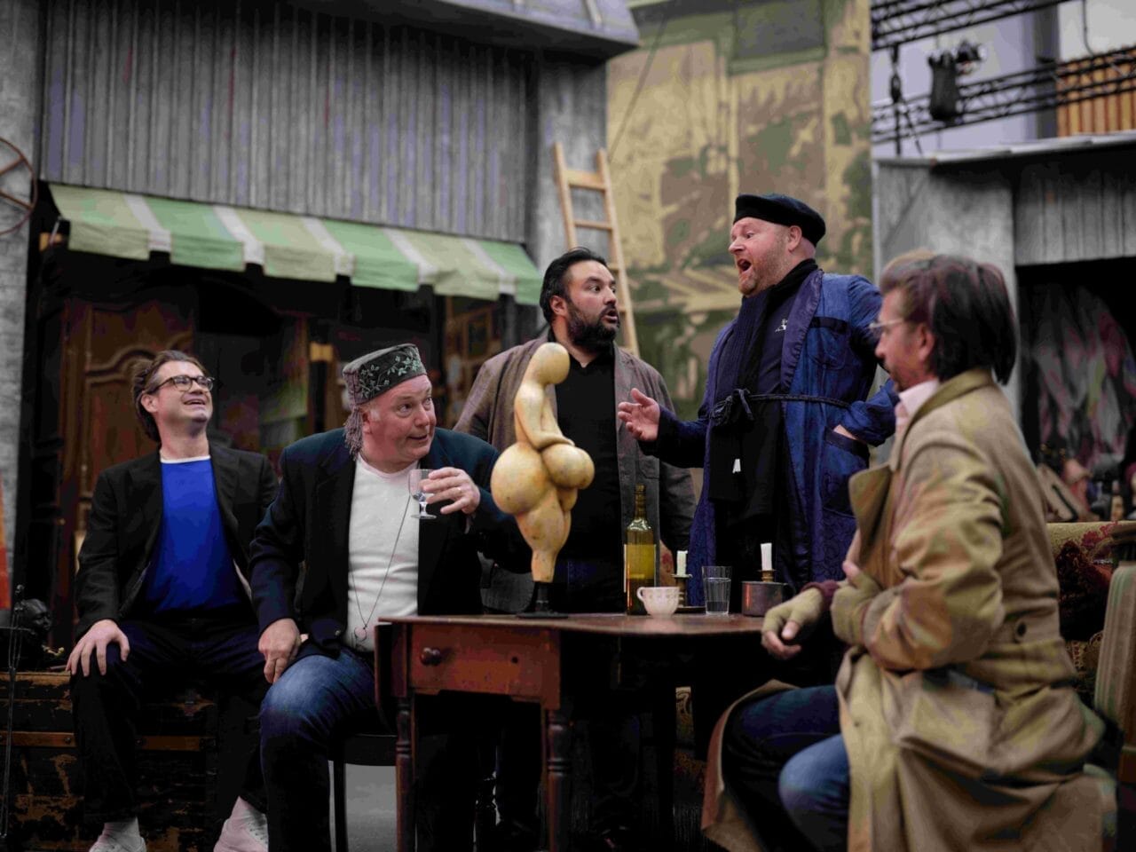 Five men sit around a table with drinks, reacting animatedly to a gourd-shaped sculpture in the center, in a setting resembling a theatrical stage or backstage area.