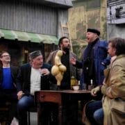 Five men sit around a table with drinks, reacting animatedly to a gourd-shaped sculpture in the center, in a setting resembling a theatrical stage or backstage area.