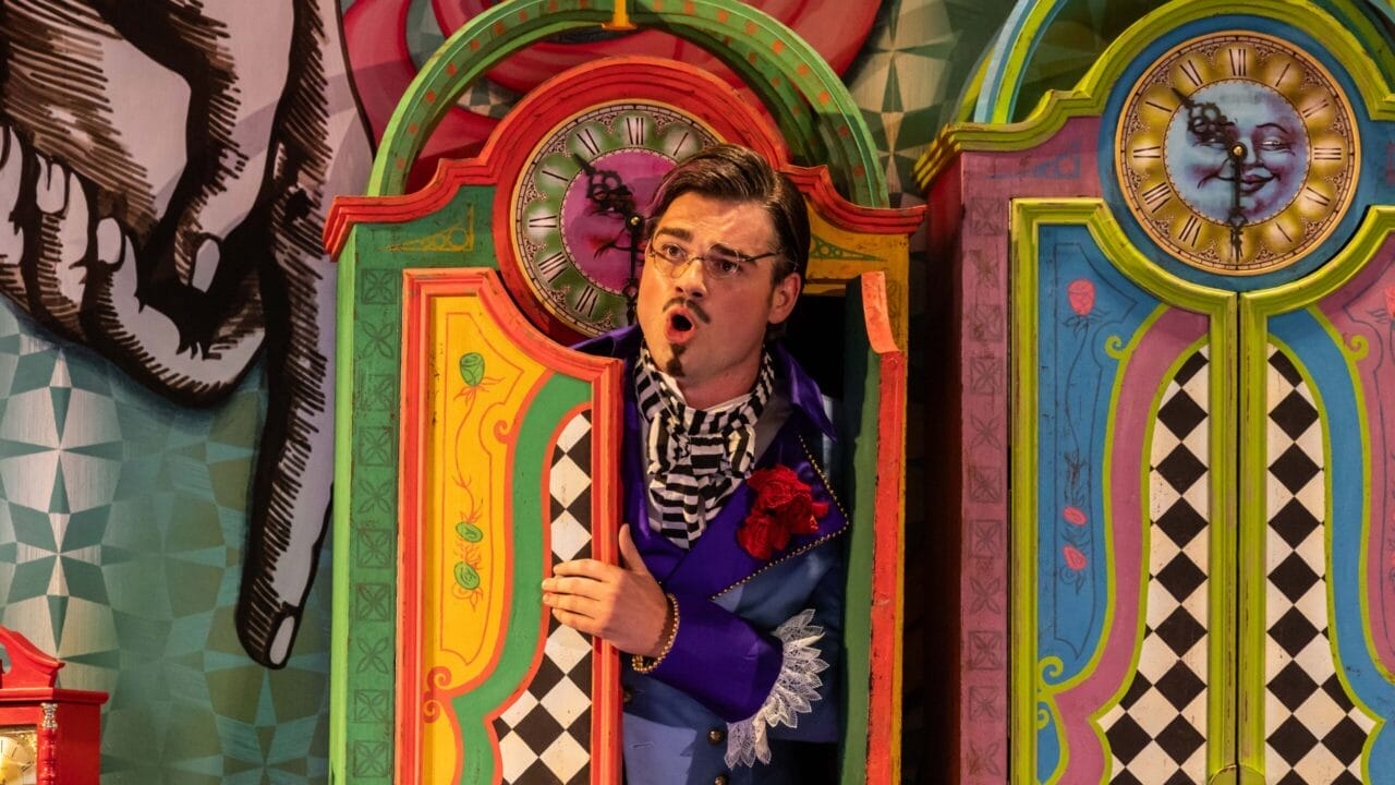 A man in colorful, theatrical clothing stands in a vibrant, painted booth with clocks and patterned doors, performing a scene from L'heure espagnole or The Bear on stage.