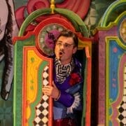 A man in colorful, theatrical clothing stands in a vibrant, painted booth with clocks and patterned doors, performing a scene from L'heure espagnole or The Bear on stage.