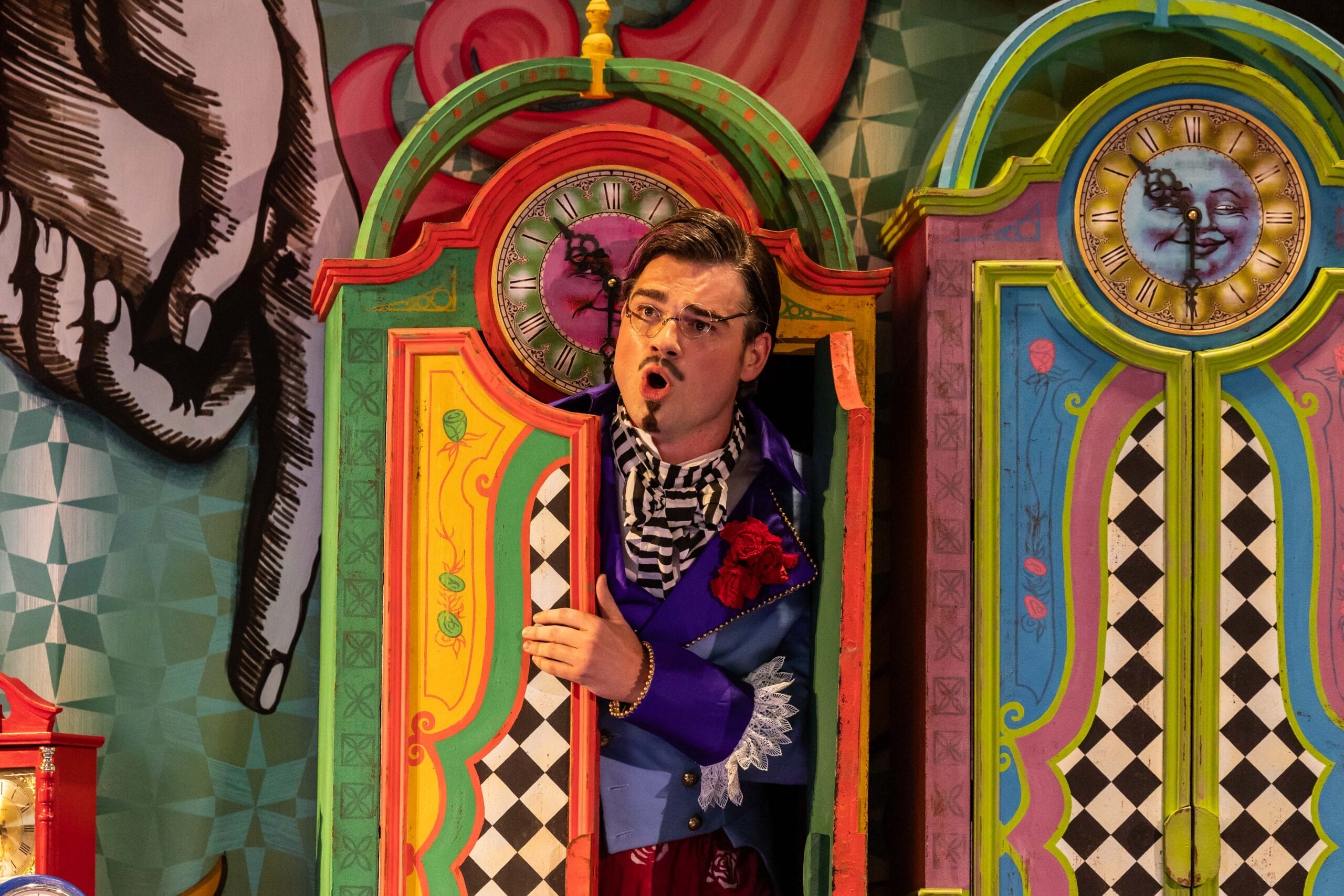 A man in colorful, theatrical clothing stands in a vibrant, painted booth with clocks and patterned doors, performing a scene from L'heure espagnole or The Bear on stage.