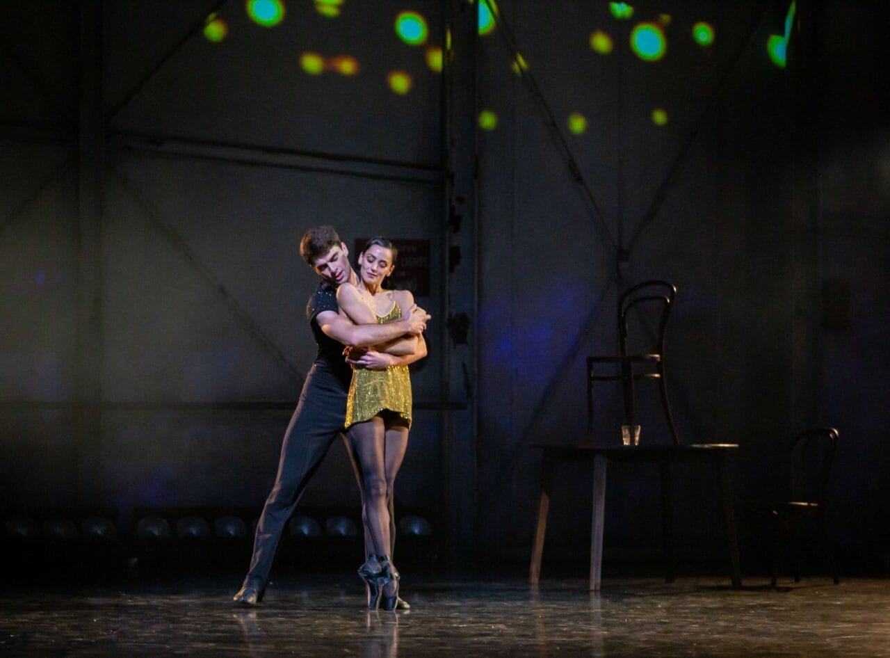 Two dancers perform an embrace on stage under spotlights, with a table, two chairs, and a glass in the background.