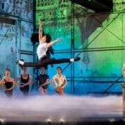 A male ballet dancer leaps mid-air on stage while other dancers and a person in a Roman-style helmet watch in the background, with colorful lighting and mist.