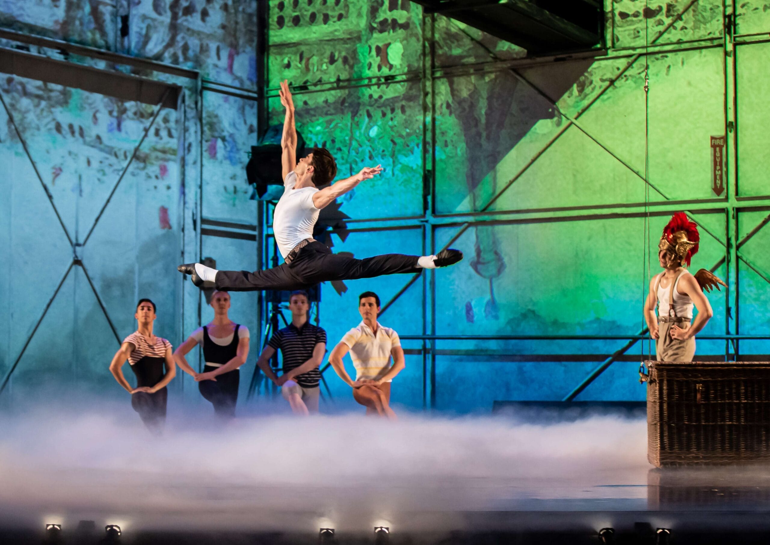 A male ballet dancer leaps mid-air on stage while other dancers and a person in a Roman-style helmet watch in the background, with colorful lighting and mist.