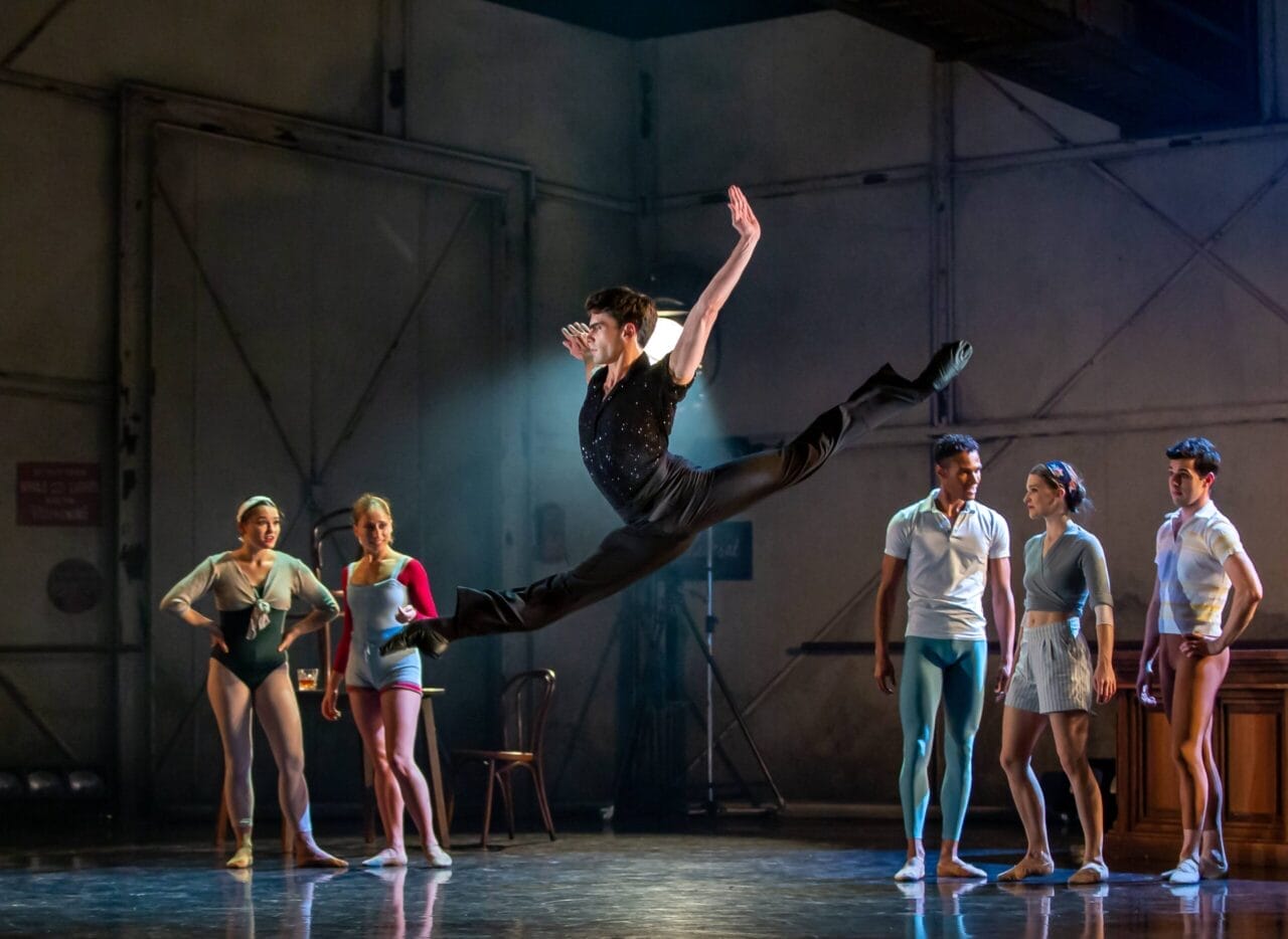 A dancer leaps mid-air with arms and legs extended while five other dancers watch on stage in a rehearsal studio setting.