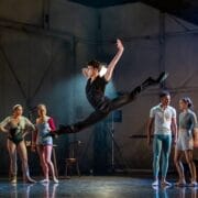 A dancer leaps mid-air with arms and legs extended while five other dancers watch on stage in a rehearsal studio setting.