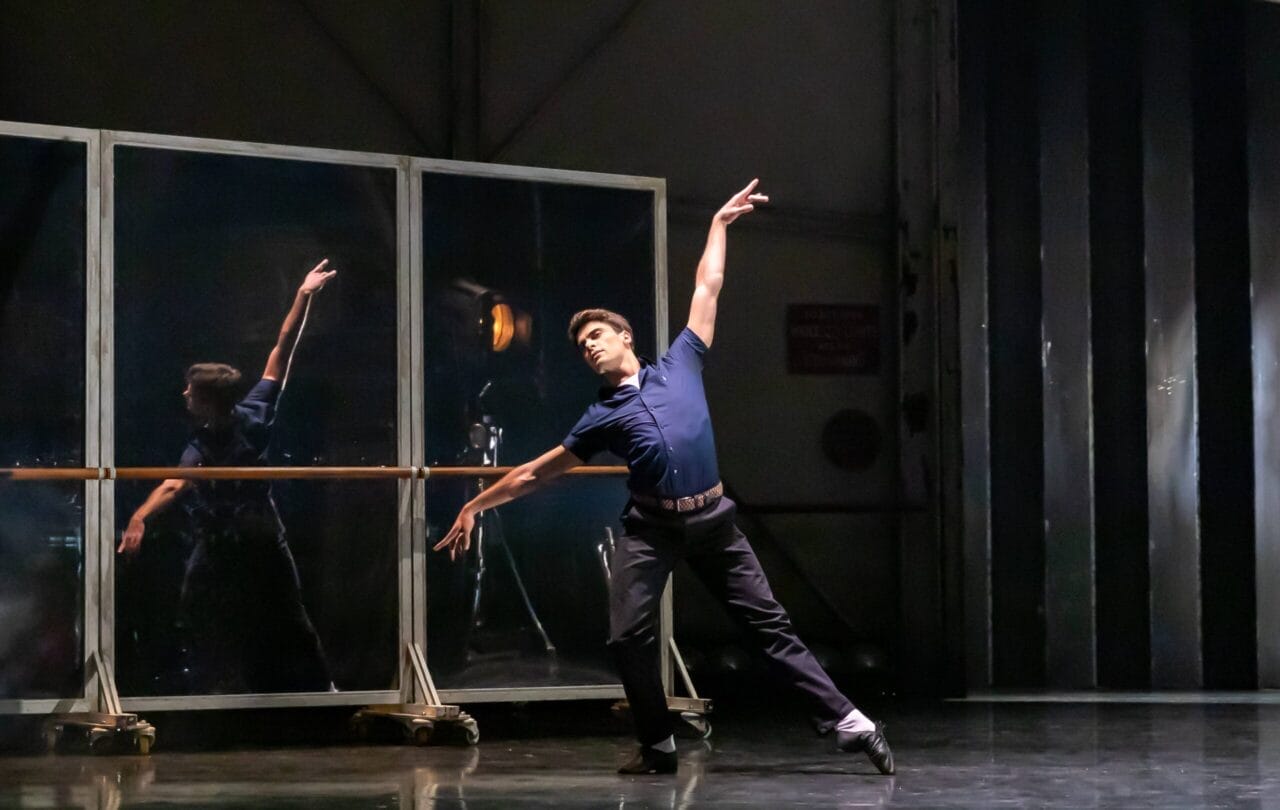 A male dancer in a navy shirt and black pants performs a pose on stage, one arm raised, in front of mirrors and metal paneling.