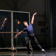A male dancer in a navy shirt and black pants performs a pose on stage, one arm raised, in front of mirrors and metal paneling.