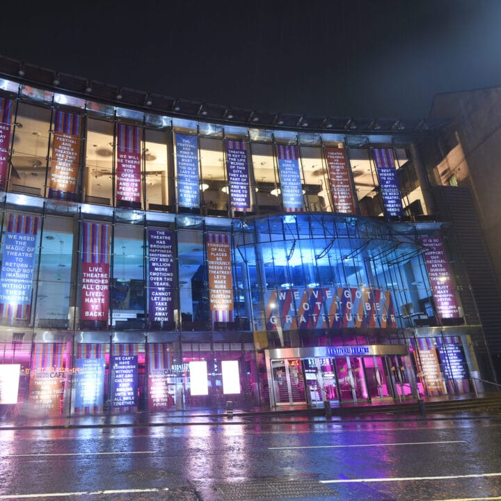 A modern glass building lit up at night with colorful illuminated signs and text covering its front facade, reflecting onto the wet street.