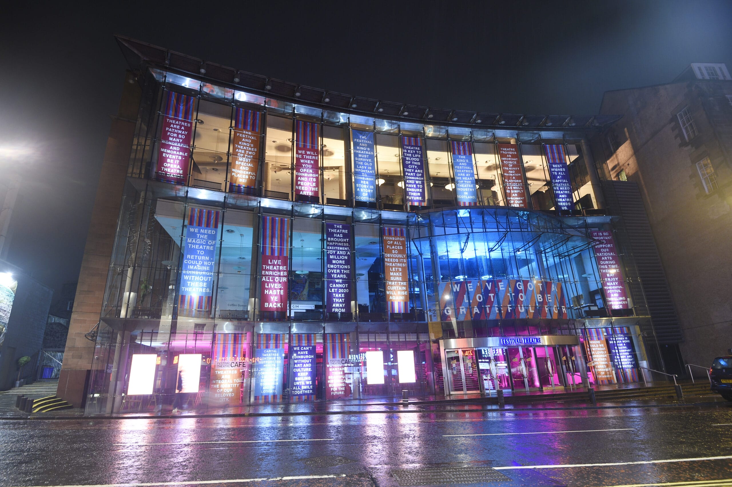 A modern glass building lit up at night with colorful illuminated signs and text covering its front facade, reflecting onto the wet street.