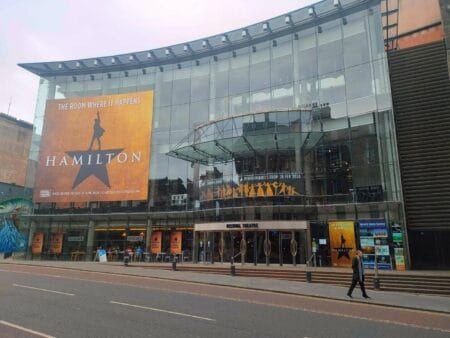 The glass-fronted Festival Theatre with "Hamilton" musical posters and signage, including a large orange banner reading "The room where it happens," reflecting onto the street outside.