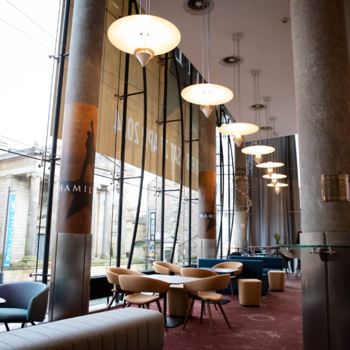 A bright and modern foyer area with round tables and chairs, large windows, hanging lights, and "Hamilton" banners, overlooking a classical building outside.