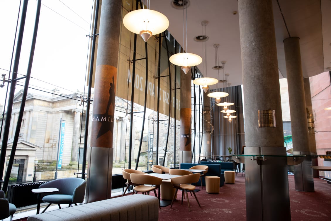 A bright and modern foyer area with round tables and chairs, large windows, hanging lights, and "Hamilton" banners, overlooking a classical building outside.