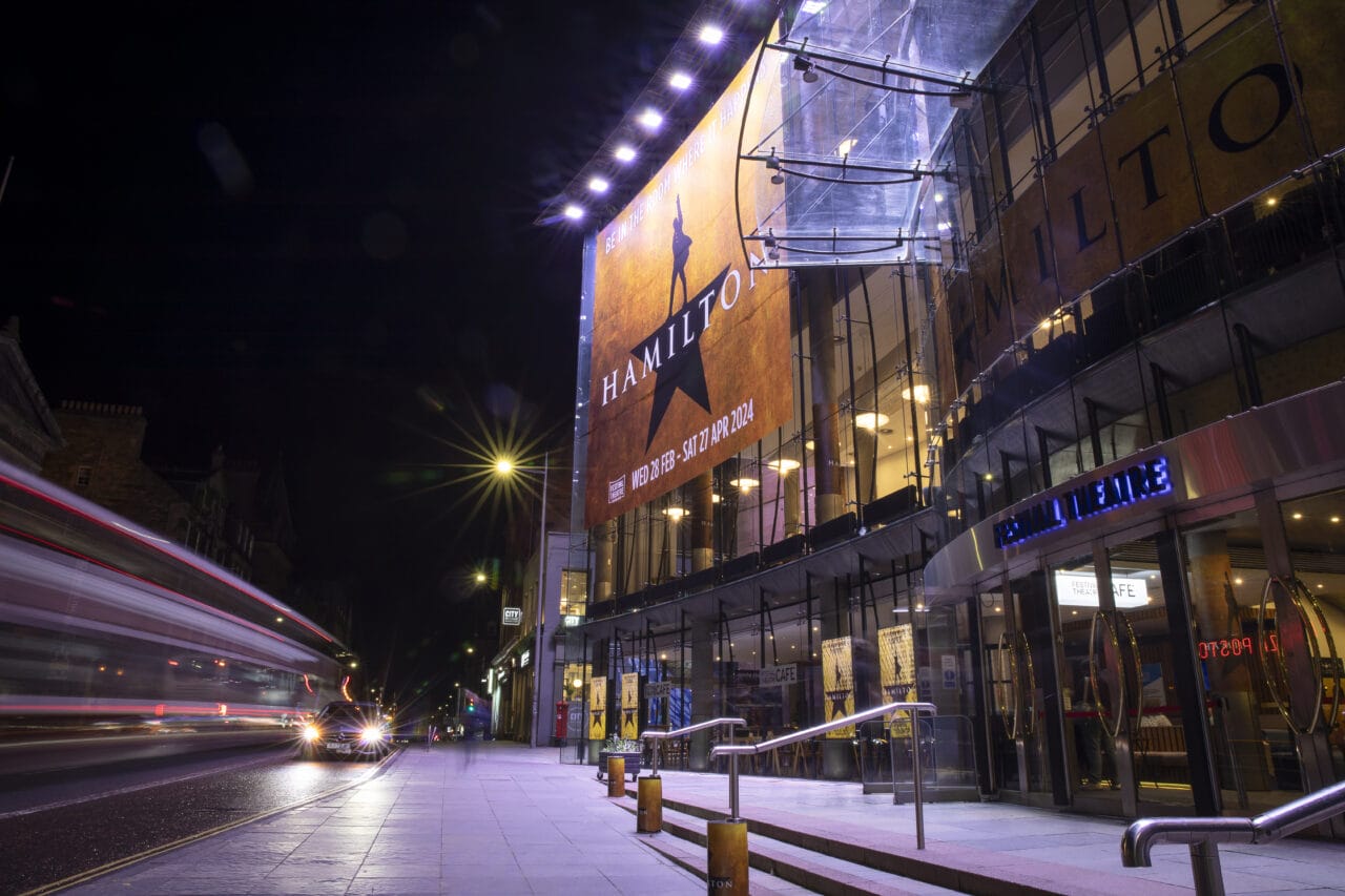 The Festival Theatre at night displays a large "Hamilton" musical banner on its glass facade, with cars and light trails visible on the street.