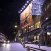 The Festival Theatre at night displays a large "Hamilton" musical banner on its glass facade, with cars and light trails visible on the street.