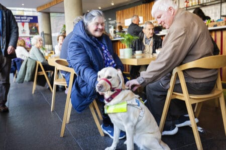 Two elderly people sit at a café table; one pets a guide dog wearing a harness while others sit and chat in the background.