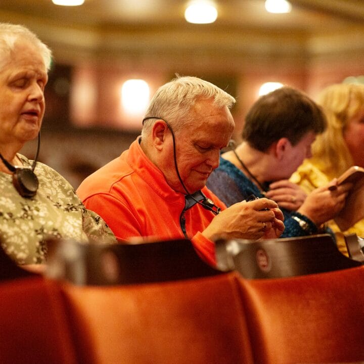 Several older adults sit in a theater audience, some using assistive listening devices and others interacting with their phones.