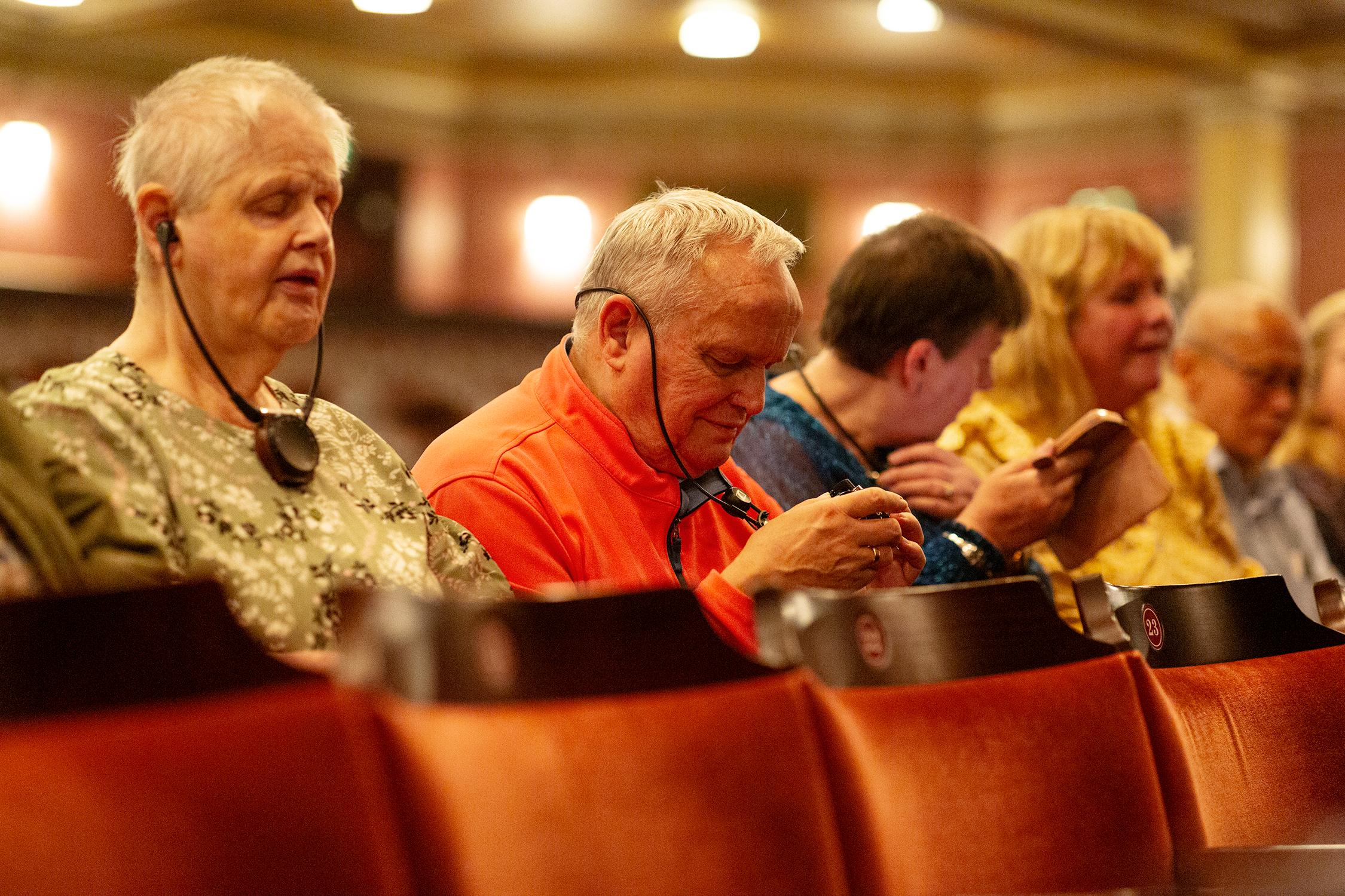 Several older adults sit in a theater audience, some using assistive listening devices and others interacting with their phones.