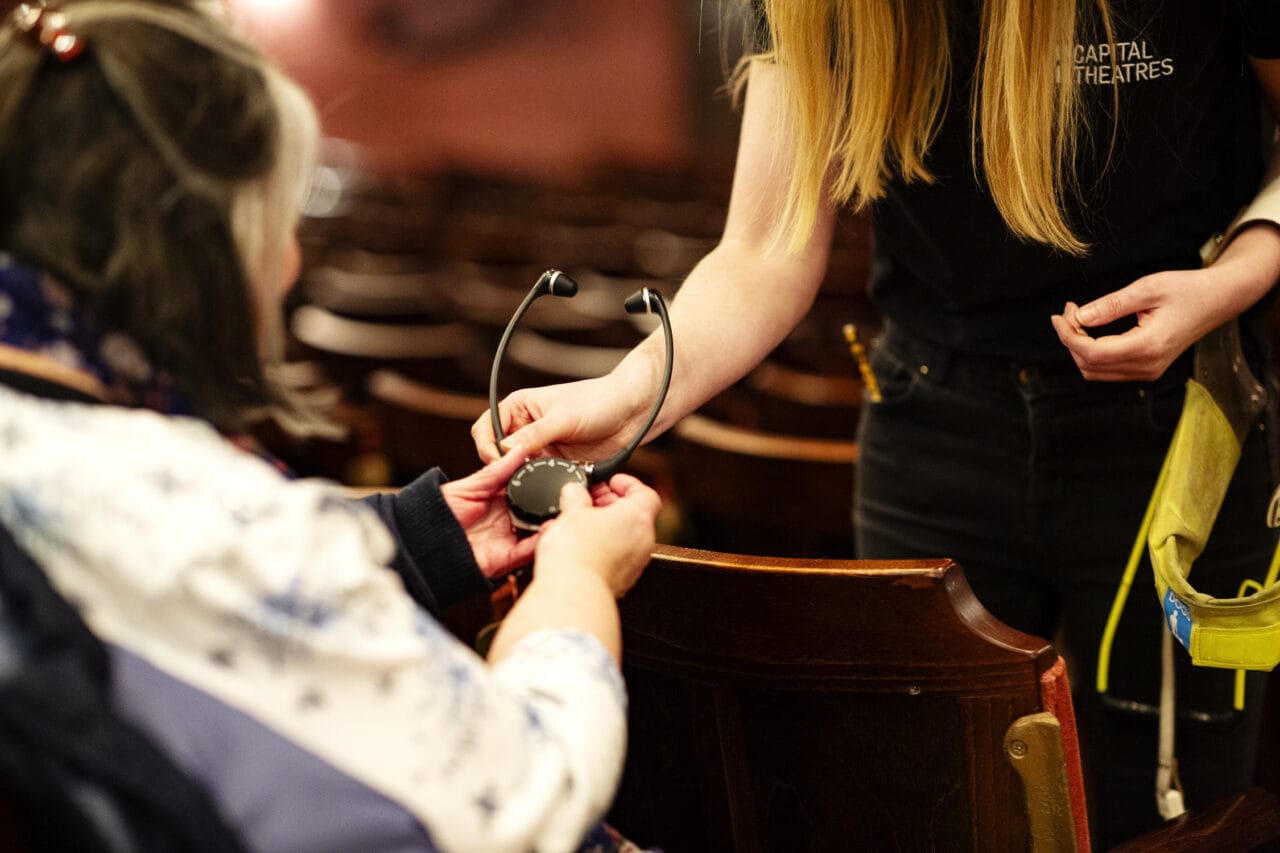 A person hands an assistive listening device to another person seated in athe theatre.