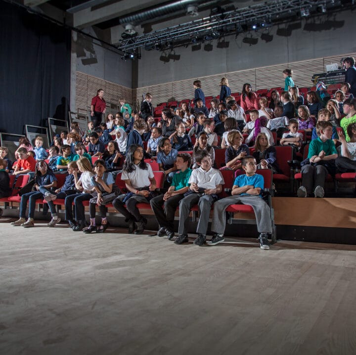 A group of children seated in an auditorium, facing forward, with an adult standing at the left side of the image.