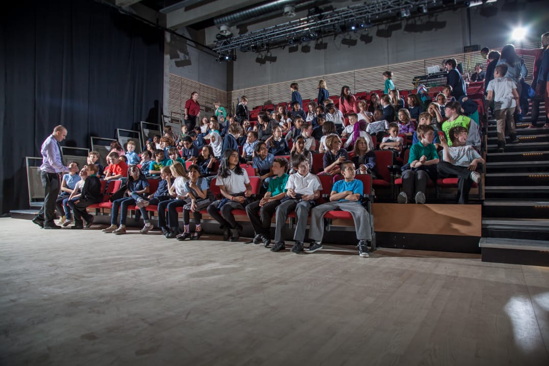 A group of children seated in an auditorium, facing forward, with an adult standing at the left side of the image.