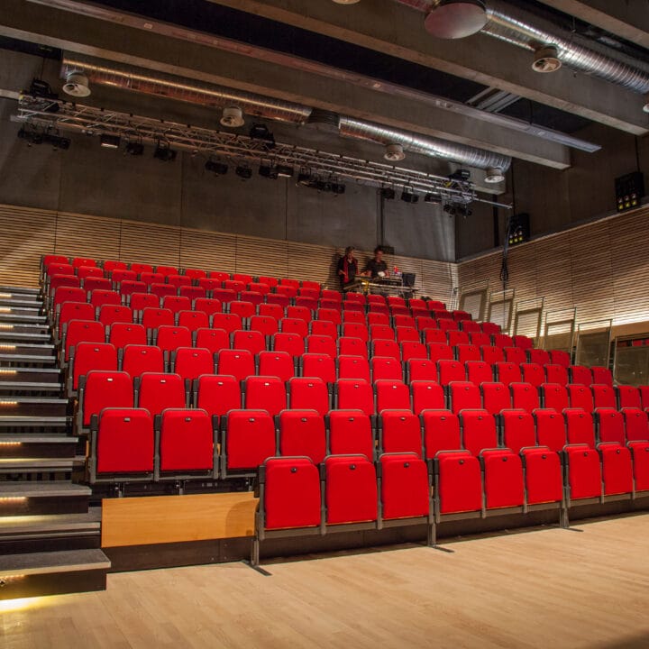 A modern auditorium with tiered rows of red seats, wooden floor, and exposed ceiling pipes, viewed from the stage area.