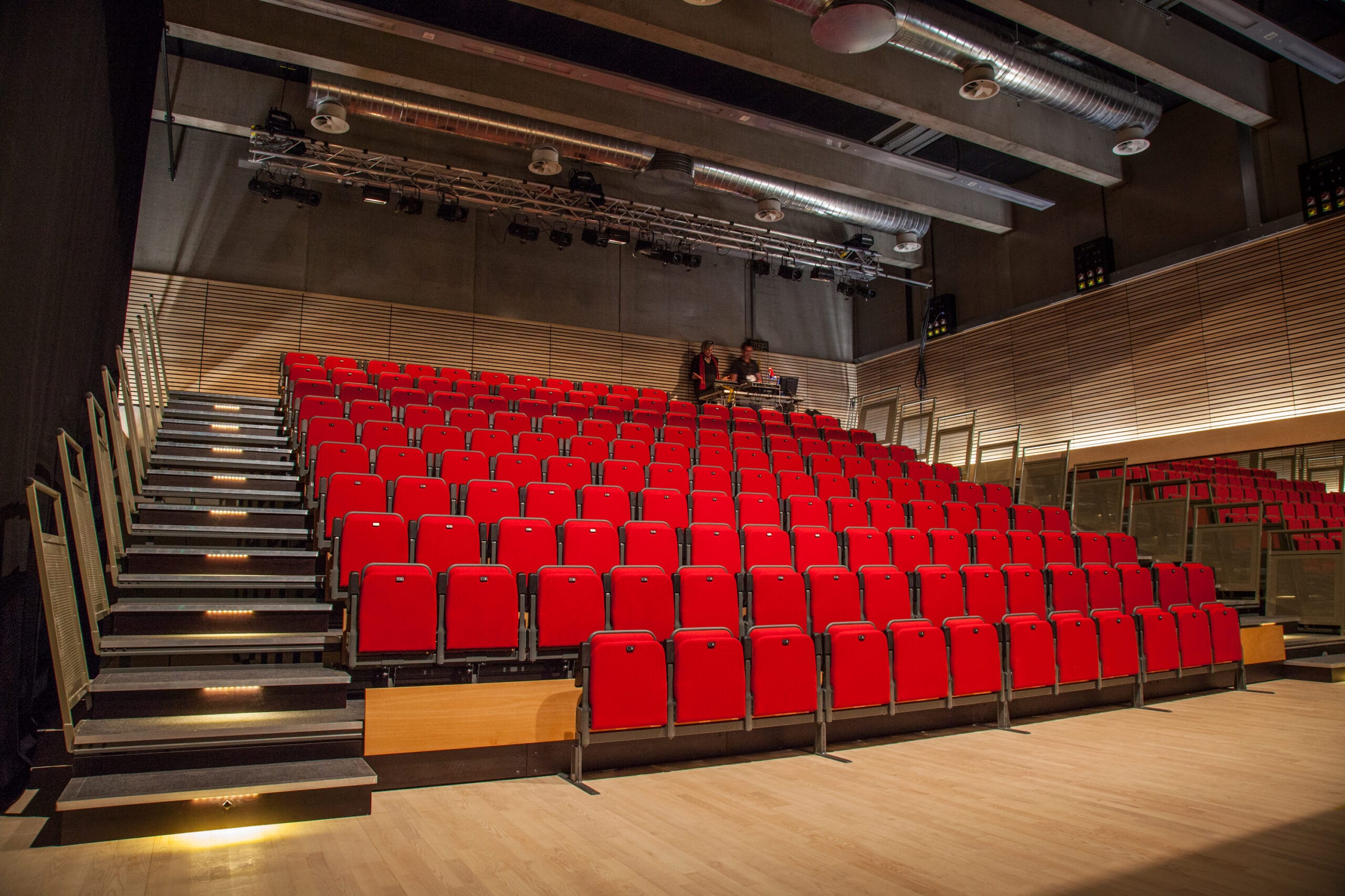 A modern auditorium with tiered rows of red seats, wooden floor, and exposed ceiling pipes, viewed from the stage area.