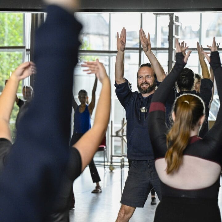 A group of people participate in a dance or exercise class, raising their arms while following an instructor at the front in a bright studio with large windows.