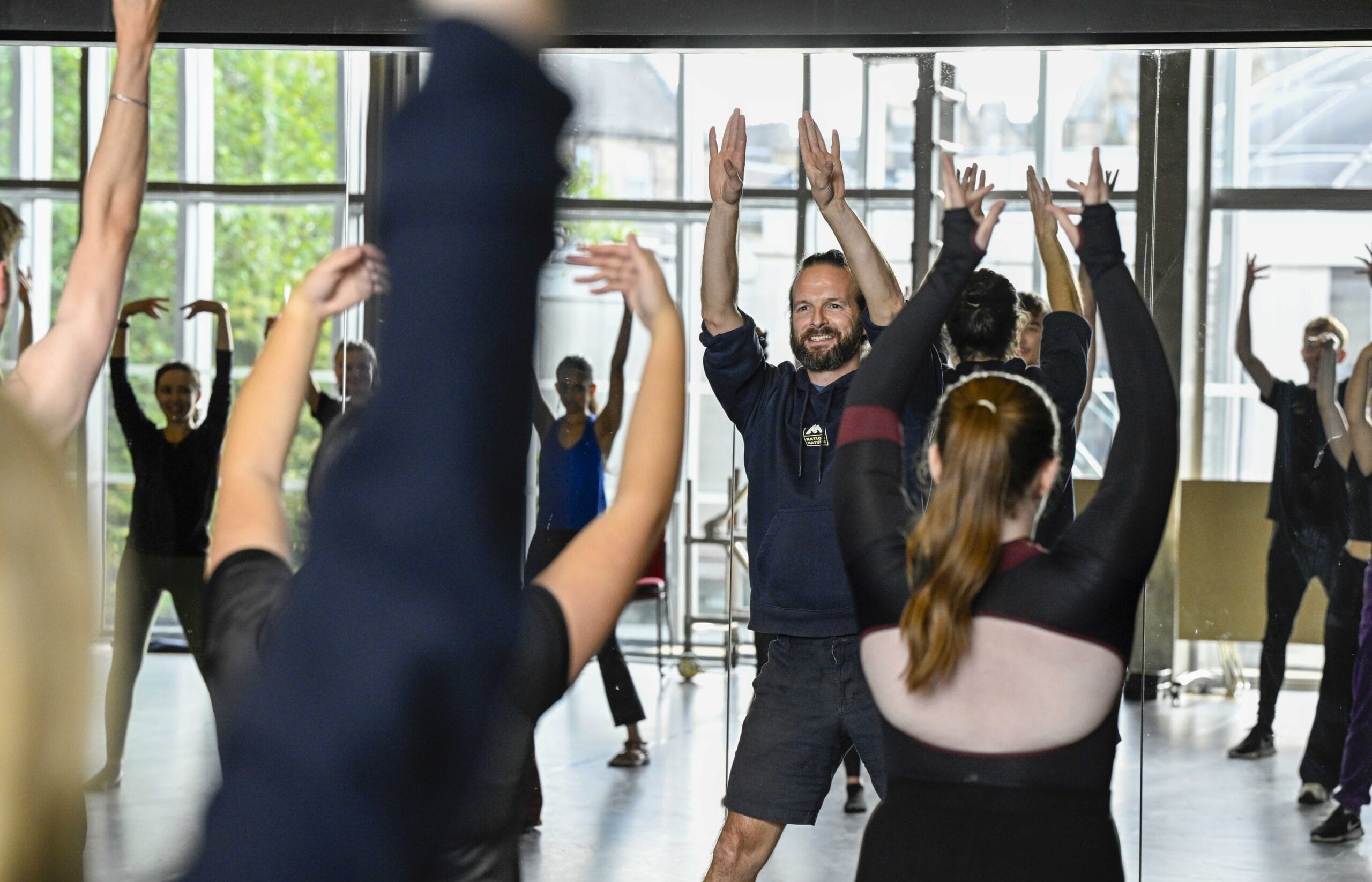 A group of people participate in a dance or exercise class, raising their arms while following an instructor at the front in a bright studio with large windows.