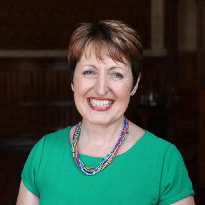 A woman with short brown hair, wearing a green top and a colorful beaded necklace, smiles while standing indoors in a wood-paneled room.