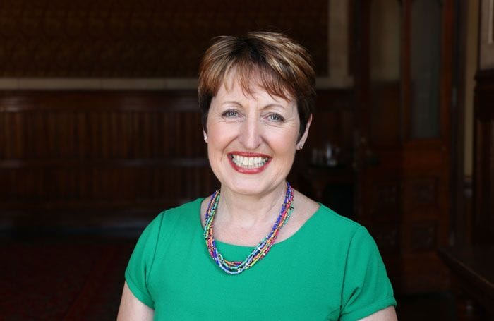 A woman with short brown hair, wearing a green top and a colorful beaded necklace, smiles while standing indoors in a wood-paneled room.