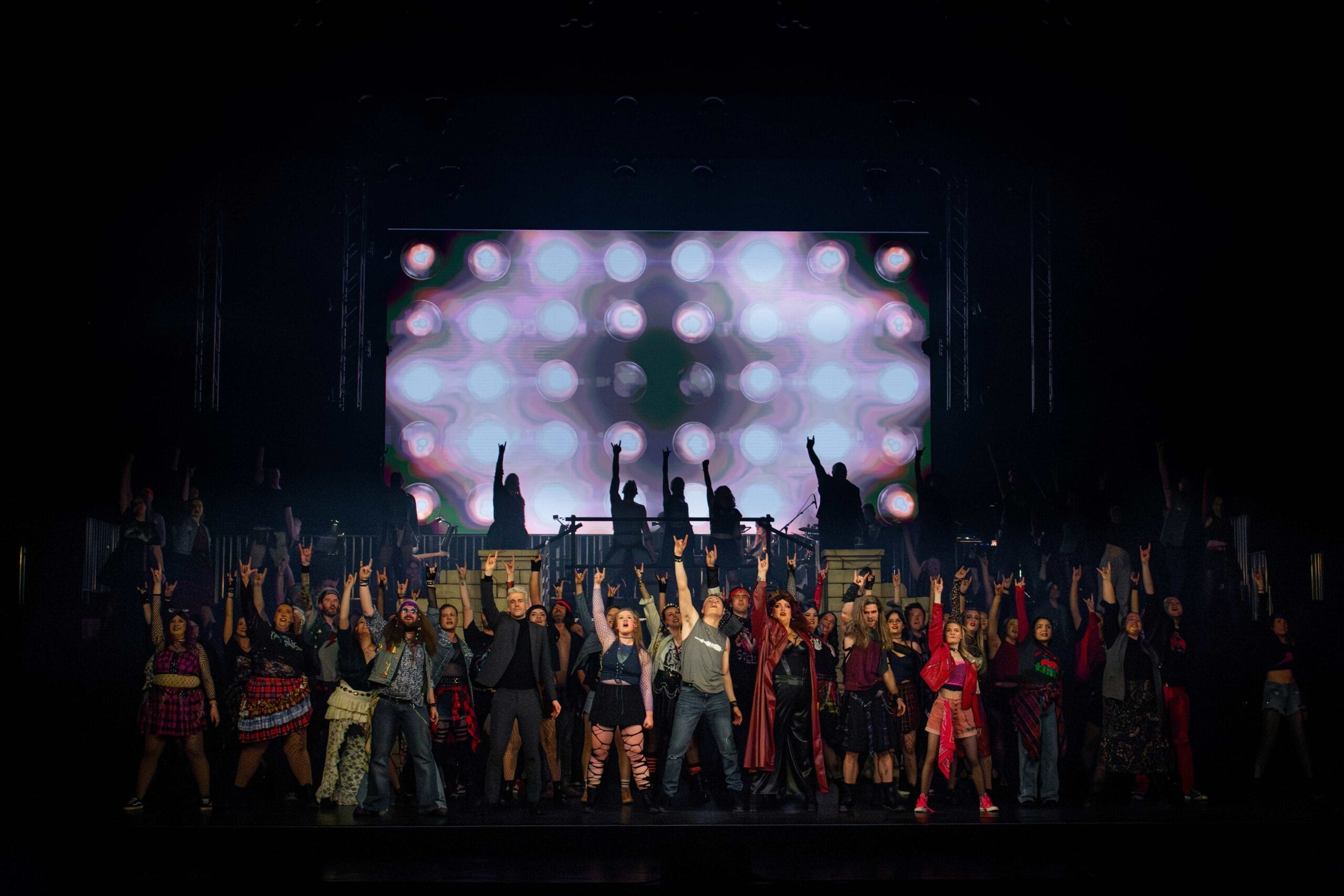 A large group of performers stand on stage in front of a backdrop of bright circular lights, posing with raised arms. The stage is dimly lit, and the group, inspired by "We Will Rock You," is dressed in a variety of colourful costumes.